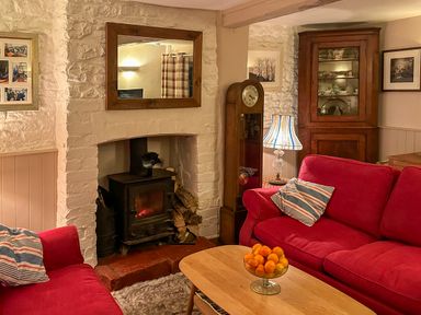 A living room with red sofas a wooden coffee table with a bowl of oranges a fireplace with a wood stove and a tall clock at Lamorna Cottage in Chipping Norton