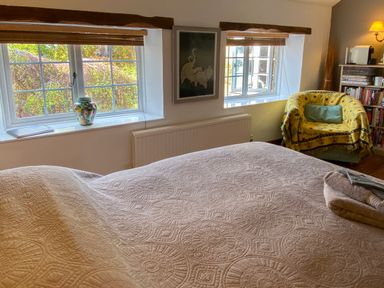 A bedroom with a bed covered with a patterned coverlet near two windows with wooden blinds and a chair with a yellow throw at Lamorna Cottage in Chipping Norton