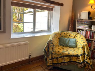 A corner with a yellow floral covered armchair a bookshelf with books and a coffee machine next to a window at Lamorna Cottage in Chipping Norton