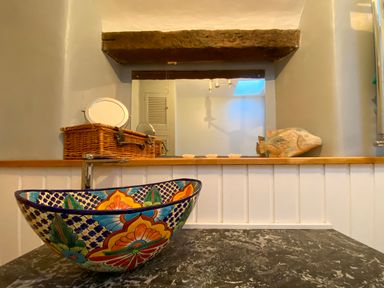 A bathroom countertop with a colorful bowl sink a wicker basket a mirror and a wooden fish decoration at Lamorna Cottage in Chipping Norton