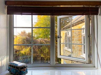 A window with one pane open showing trees and a stone building outside with a toy van on the windowsill at Lamorna Cottage in Chipping Norton