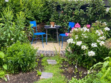 A garden with a small gravel seating area with two blue metal chairs and a table surrounded by green plants and flowers at Lamorna Cottage in Chipping Norton