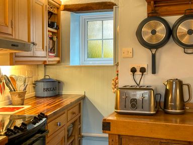A kitchen counter with utensils in a pot a bread container a toaster a kettle and pans hanging on a wall at Lamorna Cottage in Chipping Norton