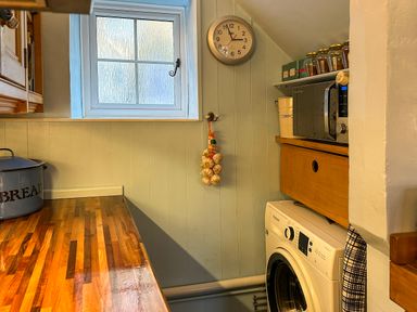 A kitchen corner with a wooden countertop a window a wall clock garlic hanging a microwave jars and a washing machine at Lamorna Cottage in Chipping Norton