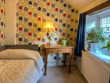 A bedroom corner with a bed a wooden side table with lamp and plant and a window with curtains and plants at Lamorna Cottage in Chipping Norton