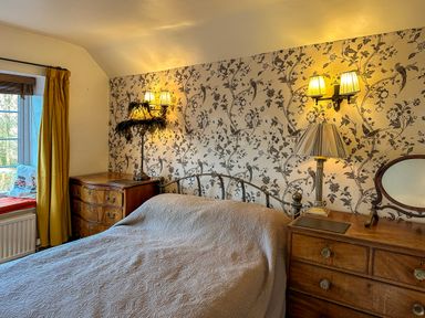 A bedroom with a bed between two wooden dressers with lamps and floral wallpaper at Lamorna Cottage in Chipping Norton