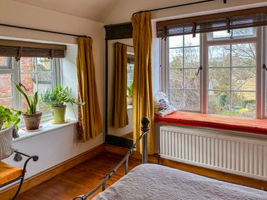 A bedroom corner with windows, potted plants on the sill, a wall mirror with mustard curtains, and a window seat with a cushion at Lamorna Cottage in Chipping Norton
