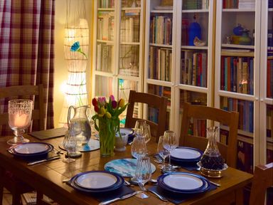 A dining table set with plates glasses and cutlery beside a bookshelf with glass doors and a lamp with floral design at Lamorna Cottage in Chipping Norton