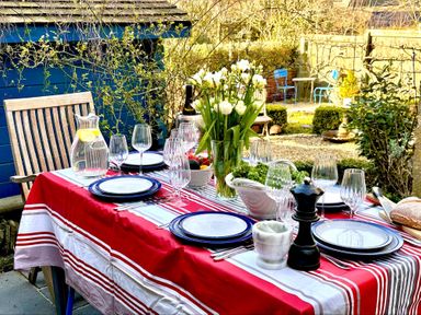 An outdoor dining table set with plates wine glasses a pitcher of water flowers and condiments in a garden