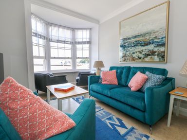 A living room with teal sofas pink cushions a wooden coffee table and large bay windows at Two @ No36 in Weymouth