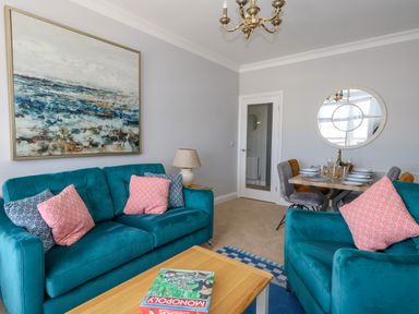 A living room with teal sofas pink and blue cushions a wooden table with a monopoly game and a dining table with plates glasses and chairs at Two @ No36 in Weymouth