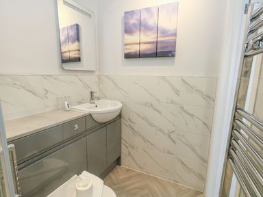 A bathroom with a sink and cabinet under, marble tiled walls, a mirror, a heated towel rail, and a beach artwork on the wall at Two @ No36 in Weymouth