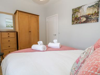 A bedroom with a bed, rolled towels, wooden wardrobe, chest of drawers with mirror, and a painting on the wall at Two @ No36 in Weymouth