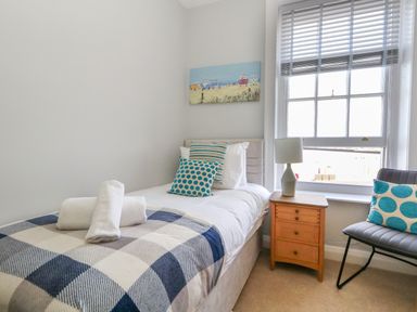 A bedroom with a single bed with pillows and folded towels a wooden nightstand with a lamp and a chair with a cushion at Two @ No36 in Weymouth