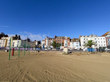 A sandy beach with volleyball poles and a row of buildings behind a road at Two @ No36 in Weymouth