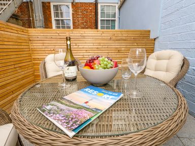 A round glass top wicker table with a bowl of fruit wine bottle magazine and four cushioned wicker chairs in a small outdoor patio at Two @ No36 in Weymouth