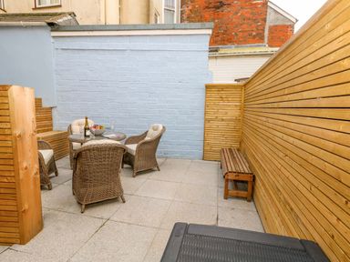An outdoor patio area with a round glass table and four wicker chairs with cushions a wooden bench and wooden panel fencing at Two @ No36 in Weymouth