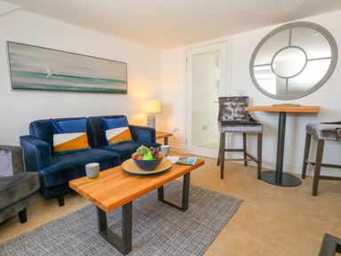 A living room with a blue sofa two grey chairs a wooden coffee table with fruit and cups a round mirror and a small dining table with two chairs at The Loft @ No36 in Weymouth