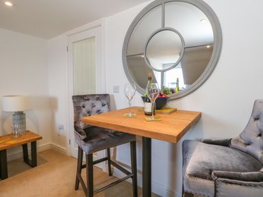 A dining area with two gray velvet chairs a wooden table with wine glasses and a bottle a round wall mirror and a side table with a lamp at The Loft @ No36 in Weymouth