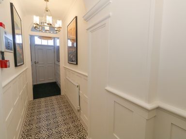 A narrow hallway with patterned floor tiles a chandelier two framed pictures and a white door at The Loft @ No36 in Weymouth