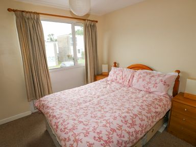 A bedroom with a double bed with pink floral bedding two wooden bedside tables with lamps and a window with beige curtains at Freshwater East in Freshwater East