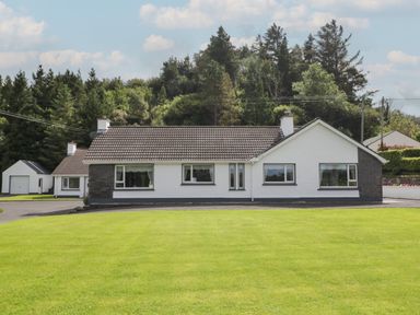A single-story white house with a tiled roof and multiple windows in front of a lawn and trees at Nelly's House in Gortahork County Donegal