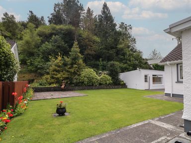 A backyard with a lawn patio flower bed and a small white building surrounded by trees at Nelly's House in Gortahork County Donegal