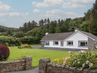 A white bungalow house with a grey roof surrounded by a green lawn and flower beds with stone walls and trees in the background at Nelly's House in Gortahork County Donegal