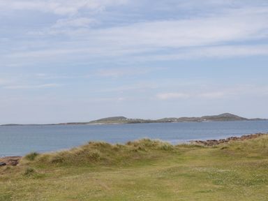 A coastal landscape with grassy dunes and a calm sea with hills in the background at Nelly's House in Gortahork County Donegal