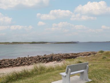 A stone bench overlooking a rocky shoreline and calm sea with small islands under a partly cloudy sky at Nelly's House in Gortahork County Donegal