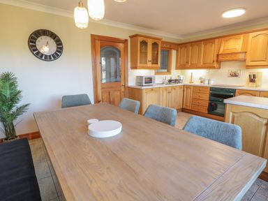 A kitchen with wooden cabinets a wooden table with plates on it blue chairs a wall clock and a plant at Nelly's House in Gortahork County Donegal