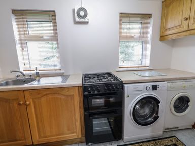 A kitchen with wooden cabinets a black stove a white washing machine a white dryer a sink and two windows at Nelly's House in Gortahork County Donegal