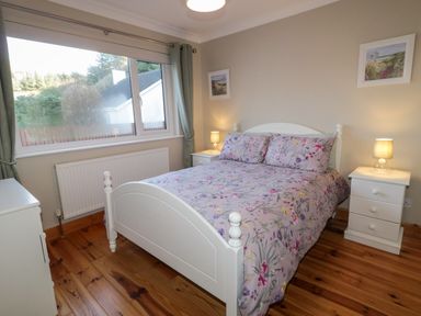 A bedroom with a white bed frame floral bedding two bedside tables with lamps wooden floor and a large window at Nellys House in Gortahork County Donegal