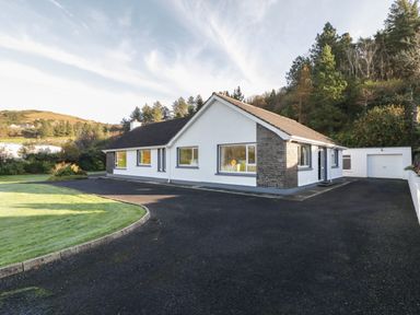 A single story house with white walls and a dark roof next to a driveway and a garage surrounded by trees at Nelly's House in Gortahork County Donegal