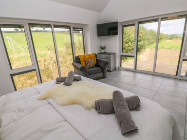 A bedroom with a bed towels a gray armchair a chest of drawers and large windows showing fields at Little Pale Near Whitland