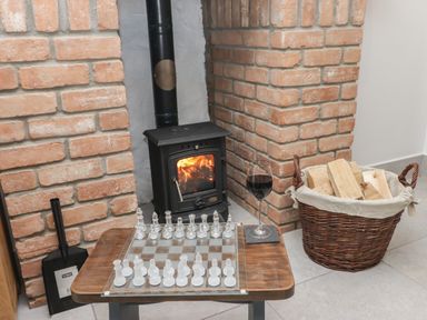 A small wood stove with fire burning next to a basket of firewood and a table with a glass chess set and a glass of red wine Near Whitland