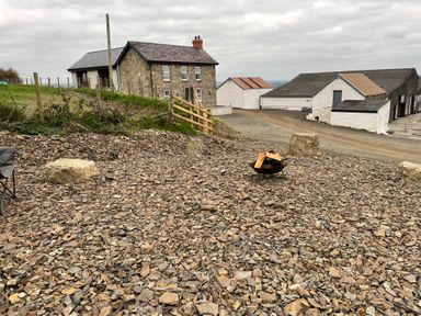 An outdoor area with scattered rocks a small fire pit with logs a stone house and farm buildings in the background at Little Pale Near Whitland