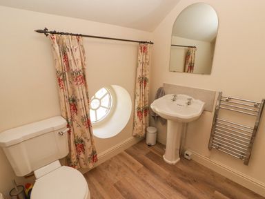 A bathroom with a toilet, pedestal sink, round window with floral curtains, mirror and towel rack at Clearbrook Lodge in Llanarthney nr Llandeilo Carmarthenshire