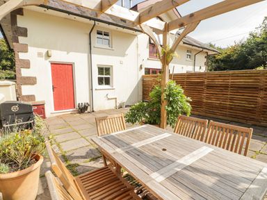 An outdoor patio with a wooden dining table and chairs under a wooden pergola near a white house with a red door and wooden fence at Clearbrook Lodge Llanarthney nr Llandeilo Carmarthenshire