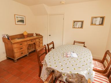 A dining room with a wooden dining table covered in a bee patterned tablecloth surrounded by wooden chairs with striped cushions and a wooden sideboard with books and a vase at Meadow Cottage in Llanarthney nr Llandeilo Carmarthenshire