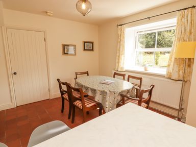 A dining room with an oval table covered with a tablecloth and six wooden chairs near a window with floral curtains at Meadow Cottage in Llanarthney nr Llandeilo Carmarthenshire