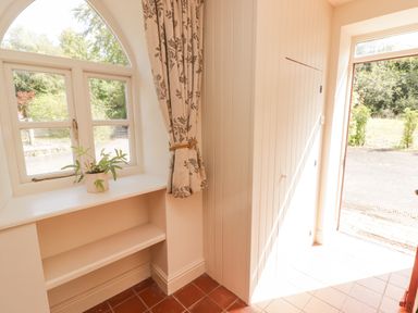 A hallway with a window, small plant on the windowsill, curtain tied back, closet door, and an open entrance door at Meadow Cottage in Llanarthney, nr Llandeilo, Carmarthenshire