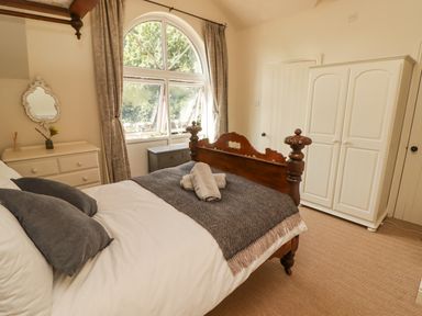A bedroom with a wooden bed with gray pillows and blanket a white dresser a white wardrobe and a large arched window at Meadow Cottage in Llanarthney nr Llandeilo Carmarthenshire