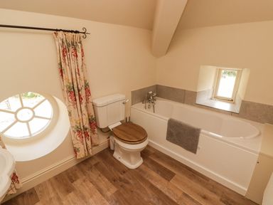 A bathroom with a bathtub a toilet with wooden seat a round window with floral curtains and a small rectangular window at Meadow Cottage in Llanarthney nr Llandeilo Carmarthenshire