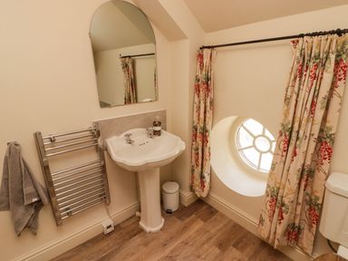 A bathroom with a pedestal sink a towel rack a round window with floral curtains and a wooden floor at Meadow Cottage in Llanarthney nr Llandeilo Carmarthenshire