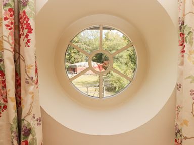 A round window with floral curtains showing a view of a barn and trees at Meadow Cottage in Llanarthney nr Llandeilo Carmarthenshire