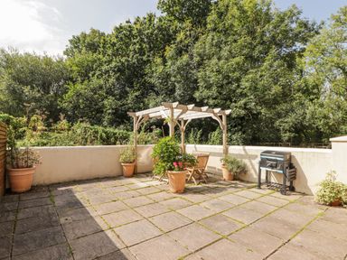 A paved patio with wooden pergola table and chairs barbecue and potted plants surrounded by trees at Meadow Cottage in Llanarthney nr Llandeilo Carmarthenshire