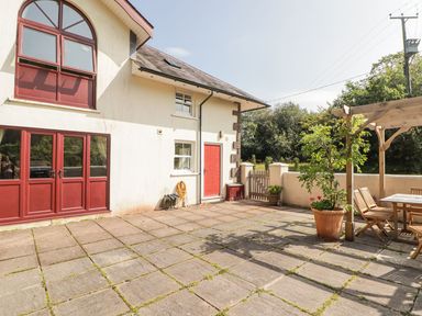 A paved patio area with red doors and windows a small wooden gate and wooden outdoor furniture under a pergola at Meadow Cottage in Llanarthney nr Llandeilo Carmarthenshire
