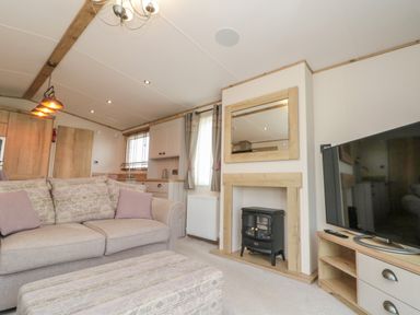 A living room with a beige sofa a patterned ottoman a small electric stove in a wooden frame a wall mirror curtains and a flat screen TV on a stand at Drake 11 in Cayton Bay