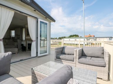 An outdoor deck with wicker chairs and a glass table next to an open door showing a living room with a dog inside at Drake 11 in Cayton Bay
